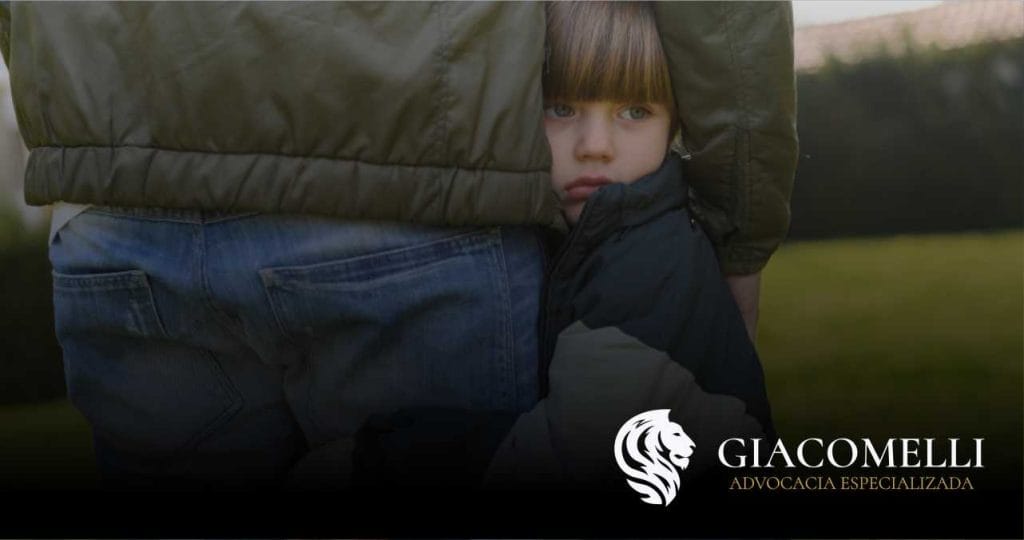 Criança abraçada à perna de um adulto (possivelmente pai), em ambiente externo; logotipo da Giacomelli Advocacia no canto inferior direito.